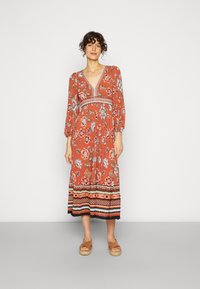 Vestuário floral de cor ferrugem com mangas longas, decote em V e bainha estampada. Apresenta uma cintura ajustada e saia fluida. Usado com sandálias castanhas.