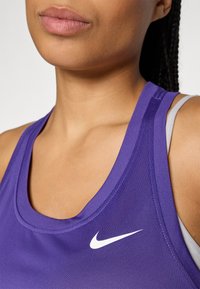 Artimas plano vaizdas, kuriame matomas žmogus, vilkintis purpurinę Nike be rankovių sportinę viršutinę dalį su balta swoosh logotipu ant krūtinės.