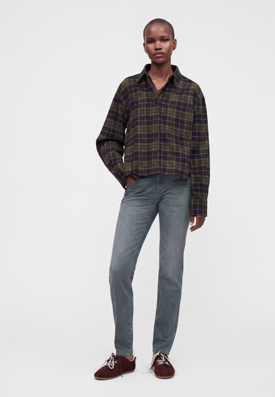 Personne debout portant une chemise à carreaux verte et noire boutonnée, un jean bleu délavé et des chaussures à lacets rouge foncé avec des lacets blancs.