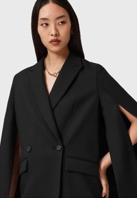 Blazer noir à double boutonnage avec une coupe ajustée, des épaules structurées et des manches ouvertes, doté de poches latérales et de accents de couture minimalistes.