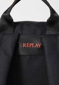 Bolso de tela negra con una superficie texturada, que presenta una prominente etiqueta roja "REPLAY" cosida en el lateral. Dos asas de transporte resistentes.