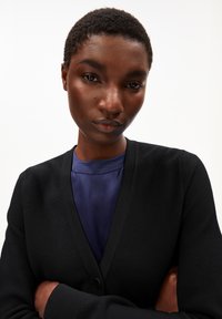 Cardigan nero con design a V, tessuto strutturato e chiusura con un solo bottone, indossato sopra una maglia blu scuro. Vista ravvicinata.