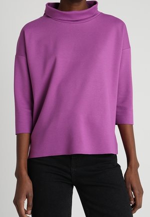 Vrouw draagt een loszittende paarse coltrui met driekwartmouwen, gecombineerd met een zwarte broek tegen een effen achtergrond.