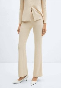 Ensemble cardigan en maille beige et pantalon évasé, tissu texturé, avec un devant boutonné, associé à des talons blancs à bout pointu.