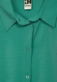 Turquoise blouse with a smooth texture, button-down front, and a classic collar. Features two buttons with a floral pattern.