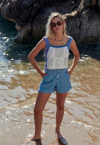 Sada crochet crop top ar ziliem akcentiem un ģeometriskām rakstām, saskaņota ar augsto vidukli džinsu šortiem pludmales vidē. Saules apgaismots ūdens fons.