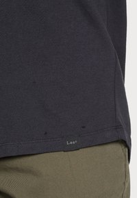 Ourlet de t-shirt gris foncé avec petite étiquette noire de la marque Lee porté sur un pantalon vert olive.
