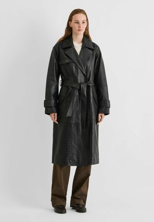 Une femme porte un long manteau en cuir noir texturé avec une ceinture nouée, assorti à un pantalon marron et à des chaussures foncées, devant un fond de studio uni.