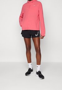 Veste rose à fermeture éclair Nike, shorts noirs avec logo blanc, chaussures de course noires, et chaussettes blanches. Comprend une capuche et des coutures contrastées.