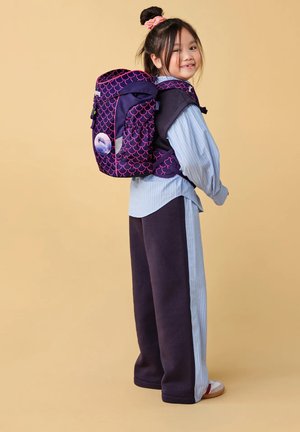 Una giovane ragazza sorridente che indossa uno zaino viola e rosa con una toppa a forma di delfino, vestita con una maglietta a righe blu e pantaloni scuri.