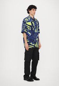 Mėlynas trumpų rankovių marškinėliai su neoninės geltonos spalvos abstrakčiu raštu, derinami su juodomis cargo kelnėmis, turinčiomis kelis kišenes ir tiesaus kirpimo.