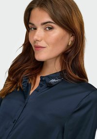 Marineblauwe shirt met een glanzende textuur, voorzien van een col met pailletten en een knopenlijst. Eenvoudige lange mouwen en een klassieke pasvorm.