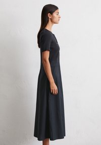 Model with long, dark hair wears a fitted, short-sleeve black dress, standing against a textured white wall, showing a profile view.