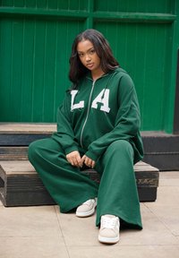 Outfit con felpa verde e pantaloni a gamba ampia, con una stampa bianca "LA", abbinato a sneakers beige, su una superficie di legno contro uno sfondo verde.