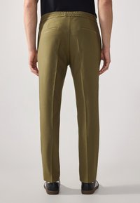 Pantaloni verde oliva con vestibilità comoda, vita elasticizzata e due tasche posteriori, abbinati a scarpe scure con strisce bianche.
