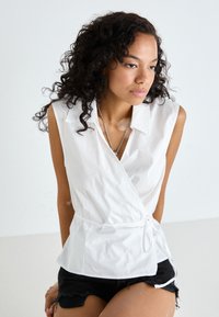 White wrap blouse with short sleeves, collar, and waist tie detail. Worn over black distressed shorts, featuring a smooth texture.