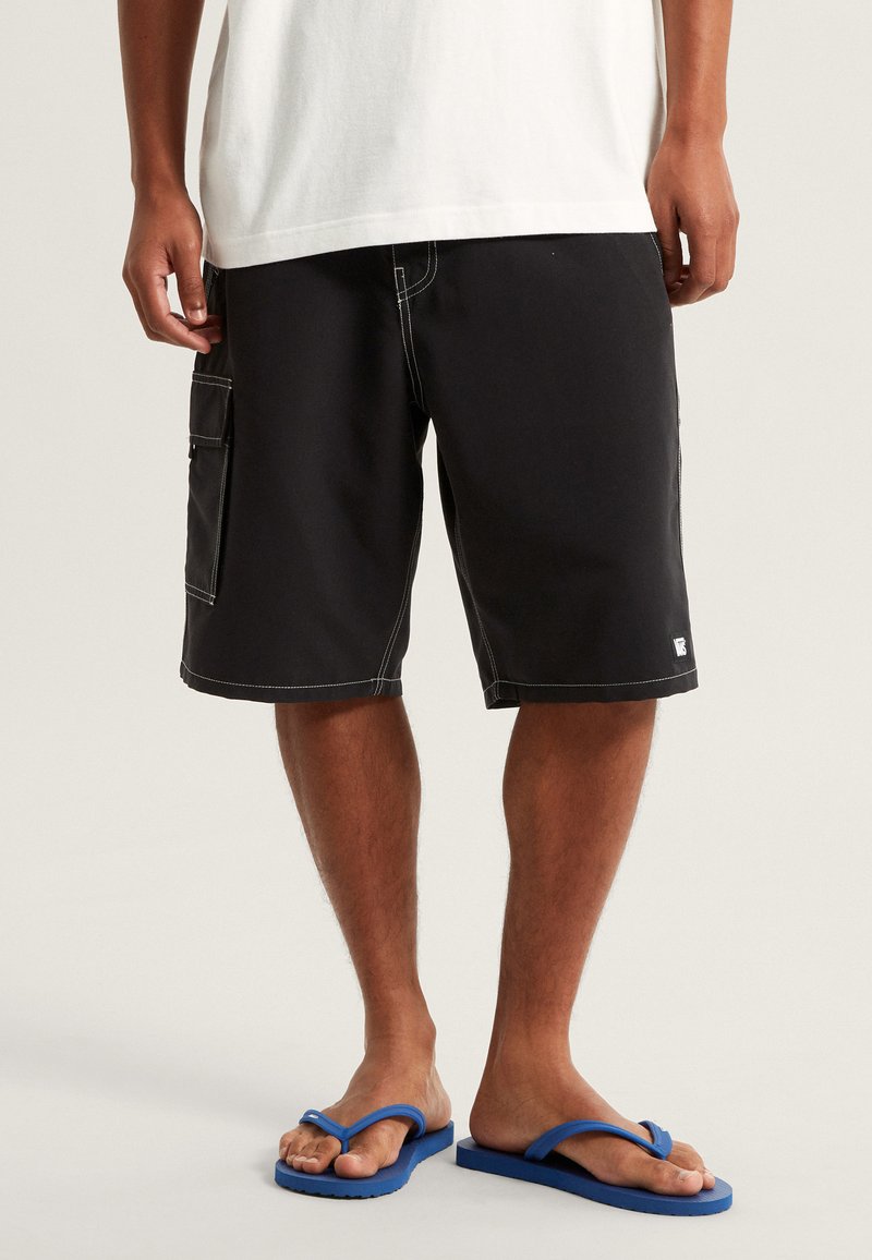 Pantaloni cargo neri con cuciture bianche, tasca laterale e patch con logo. Abbinati a ciabatte blu. Design semplice e casual, adatto per il clima caldo.