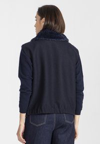 Kobieta ubrana w ciemnonawigacyjny żakiet z pluszowym kołnierzem i długimi rękawami, w połączeniu z ciemnoniebieskimi dżinsami, zwrócona plecami do aparatu.