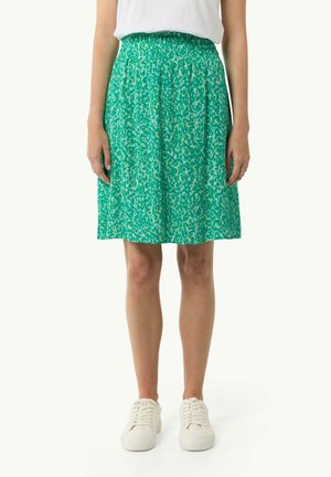 Femme portant une jupe verte à motifs longueur genoux avec une taille élastique et des baskets blanches, debout devant un fond uni.