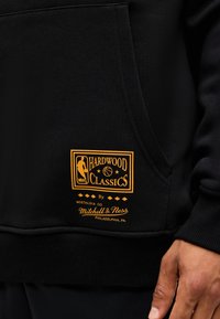 Μαύρη τσέπη φούτερ με κίτρινο λογότυπο NBA Hardwood Classics και κείμενο Mitchell & Ness Philadelphia πάνω από το χέρι ενός ατόμου που ξεκουράζεται κοντά.