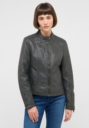 Frau mit kurzem braunem Haar, die eine graue Lederjacke mit Reißverschluss und blaue Jeans trägt, steht vor einem neutralen hellen Hintergrund.