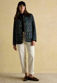 Jachetă matlasată de culoare verde închis cu un model în diamante, adornită cu butoane aurii și margini maro, purtată peste un top negru și pantaloni albi.