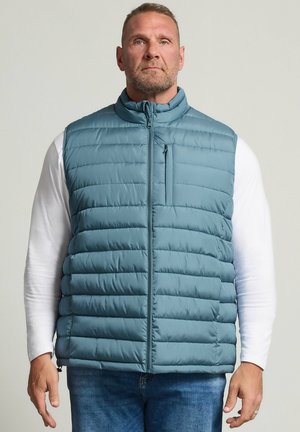 Homme portant une veste matelassée bleue sans manches par-dessus une chemise blanche à manches longues et un jean bleu, debout devant un fond clair uni.