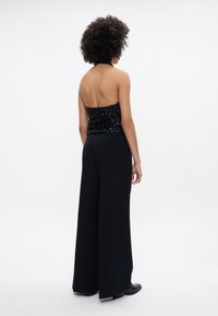Top halter preto com lantejoulas, combinado com calças largas pretas. O conjunto apresenta um corpete justo e uma parte inferior fluida, enfatizando a elegância e a estrutura.