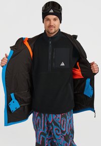Chaqueta de forro polar negra con bolsillo con cremallera, chaqueta con capucha marrón, forro en azul brillante y naranja, y pantalones estampados en espirales rosas y azules.