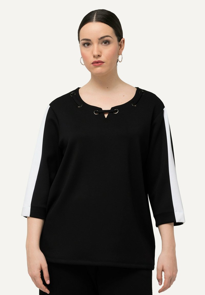Czarna bluza z dekoltem w kształcie klucza, z koronkowymi detalami i białymi paskami po bokach. Posiada rękawy o długości trzech czwartych oraz gładką teksturę.