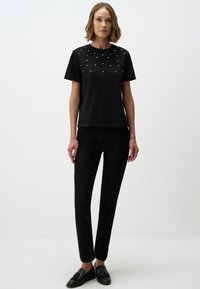 Zwarte korte mouwen t-shirt met parelachtige studs op de borst, gecombineerd met zwarte slim-fit broek en zwarte instappers. Gladde stoftextuur.