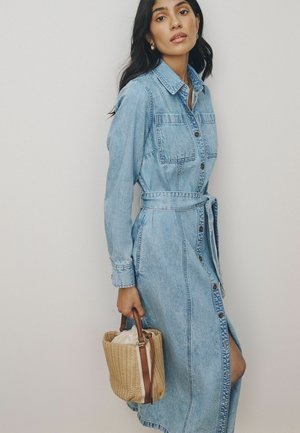 Woman with long dark hair wears a belted, button-up denim dress and holds a small woven straw handbag with leather handles.
