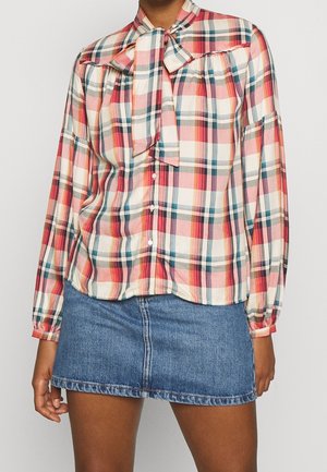 Vrouw draagt een blouse met lange mouwen in roze, rood en beige ruitjespatroon met strik bij de hals en knopen aan de voorkant, gecombineerd met een blauwe denim minirok.