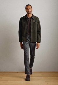 Hombre caminando en interiores con chaqueta de botón verde oscuro, polo oscuro, pantalones grises y mocasines oscuros sobre un suelo de madera.