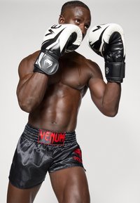 Boxeador com luvas pretas e brancas, vestindo calções de cetim pretos com um logo vermelho "Venum". Construcção musculosa, braços erguidos numa posição de boxe.