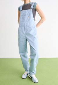 Hellblaue Denim-Latzhose mit verstellbaren Trägern, einer Brusttasche und seitlichen Knöpfen, kombiniert mit einem enganliegenden grauen T-Shirt und Turnschuhen.