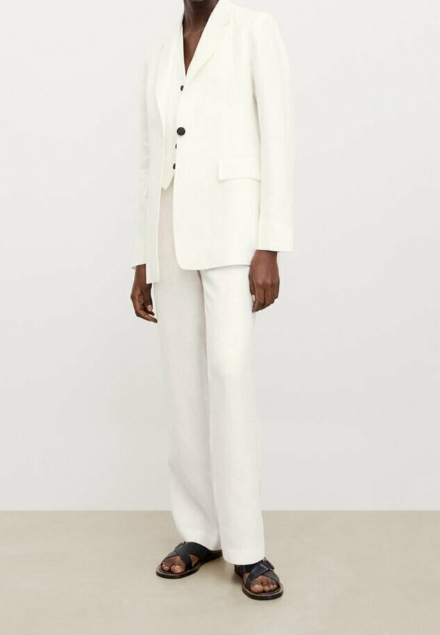 Vaaleanvärinen pellavapuku, jossa on istuva bleiseri, jossa on kaksi nappia ja sivutaskut, yhdistettynä leveälahkeisiin housuihin ja mustiin sandaaleihin.