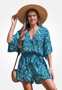Bunt gemusterter Jumpsuit mit V-Ausschnitt, weiten Ärmeln und gebundenem Taillenband. Getragen mit einem Strohhut und einer runden geflochtenen Tasche.