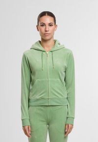 Zöld velúr cipzáras kapucnis pulóver elülső zsebbel, állítható húzózsinórral és aranyszínű fém kiegészítőkkel. Puha tapintású, hímzett logó részletekkel.