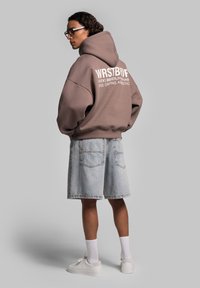 Brauner, oversized Hoodie mit weißem Text auf dem Rücken, kombiniert mit hellblauen Jeansshorts und weißen Sneakern. Glattes Material, lockere Passform.