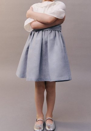 Jupe trapèze gris clair avec plis, associée à un haut blanc à manches ballon. Des ballerines argentées complètent la tenue. Fond uni.