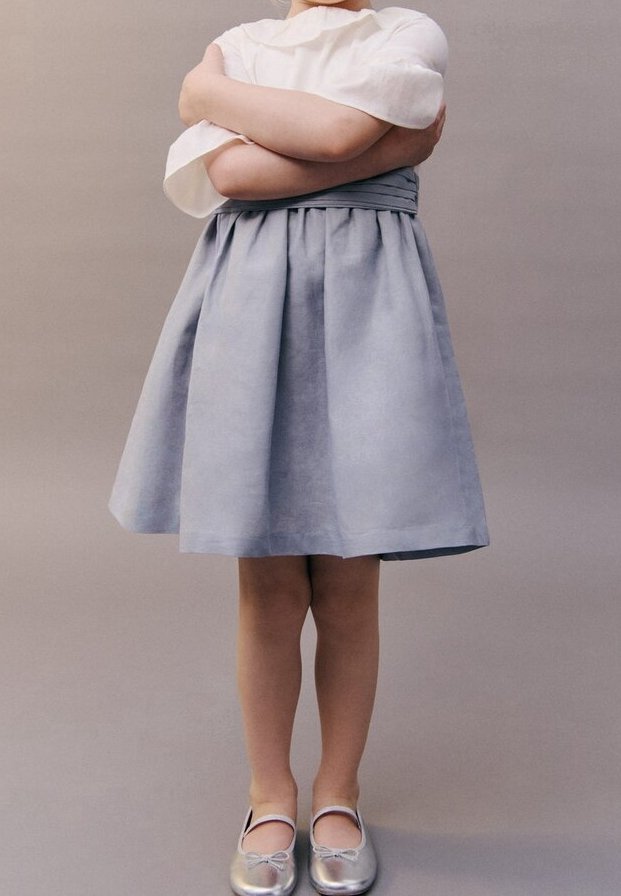 Jupe trapèze gris clair avec plis, associée à un haut blanc à manches ballon. Des ballerines argentées complètent la tenue. Fond uni.