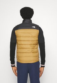 Vadderad väst i svart och brunt, med hög krage och sömnadsdesign. Logotyper på vänster axel, matchad med en svart långärmad tröja.