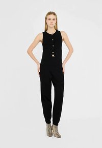 Zwart mouwloos topje met knopen, gecombineerd met zwarte broek. Model draagt hooggehakte laarzen met patroon. Eenvoudig ontwerp, aansluitende pasvorm.