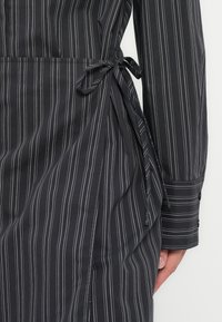 Μαύρο και άσπρο φόρεμα με ρίγες και μακριά μανίκια, με λεπτομέρεια δέσιμο στη μέση. Το υλικό φαίνεται λείο και ελαφρύ.