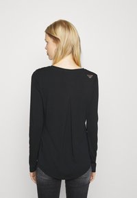 Zwarte langetermijn top met een ronde hals, een rugplooidesign en een subtiele logo-accent nabij de schouder, gemaakt van zachte stof.