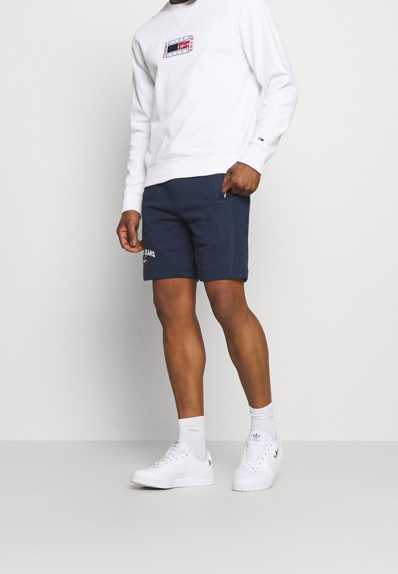 Felpa bianca con logo, pantaloni corti blu navy e sneakers bianche. I pantaloni corti presentano tasche laterali e dettagli ricamati.