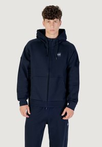 Marineblaue Zip-Hoodie aus weichem Stoff, mit Kapuze, Fronttaschen und einem Logopatch auf der Brust. Mattes Finish mit gerippten Bündchen.