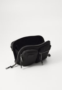 Sac en nylon noir en bandoulière avec deux compartiments zippés, extérieur texturé, quincaillerie argentée et logo à l'avant.
