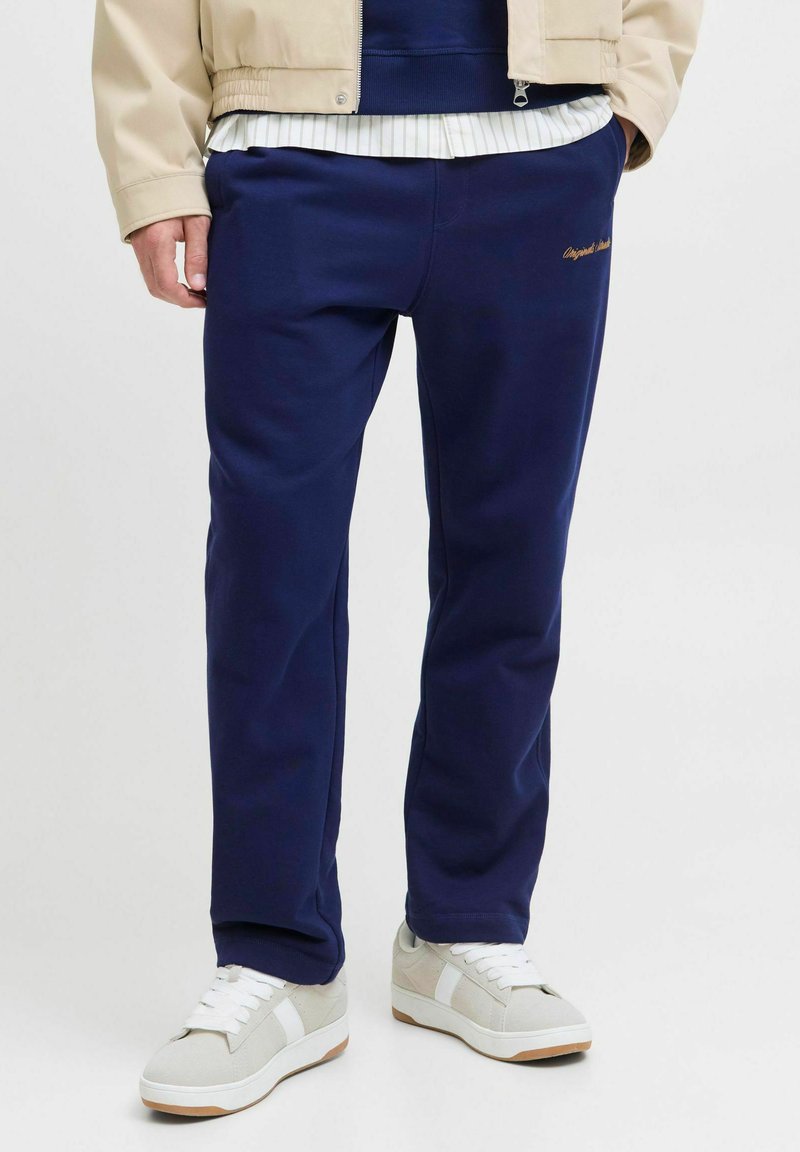 Marineblå sweatpants i et blødt stof, med et broderet logo i guld. Satsen er kombineret med hvide sneakers og en beige bomberjakke.
